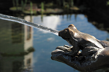 Wasserstrahl aus einer Frosch-Brunnenfigur vor ruhigem Teich im Hintergrund